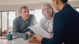 En ældre mand og kvinde sidder ved et bord med to krus og lytter opmærksomt til en person overfor dem, der holder nogle papirer i hånden – måske diskuterer de, hvad de skal gøre, hvis de er utilfredse med banken, i dette lyse og hyggelige rum.