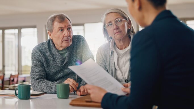 En ældre mand og kvinde sidder ved et bord med to krus og lytter opmærksomt til en person overfor dem, der holder nogle papirer i hånden – måske diskuterer de, hvad de skal gøre, hvis de er utilfredse med banken, i dette lyse og hyggelige rum.
