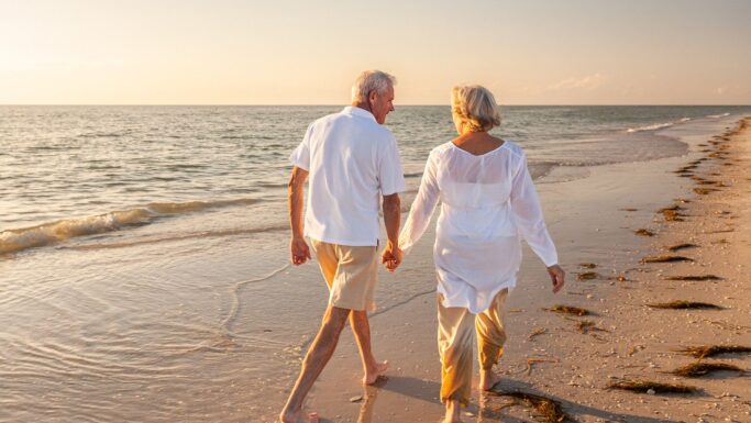 Et ældre ægtepar går hånd i hånd langs en sandstrand ved solnedgang, iført let tøj og smilende, mens de nyder den fredfyldte kyst og tænker tilbage på deres liv sammen, nu hvor de er på vej på pension.