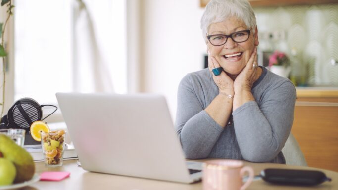 En smilende ældre kvinde med kort gråt hår og briller sidder ved et skrivebord med en bærbar computer, en kop og noget frugt. Hun har hænderne lagt mod ansigtet og ser ind i kameraet i et lyst, hyggeligt rum – hun nyder livet som en af de mange pensionister, der har fundet måder at forblive aktive og engagerede på.