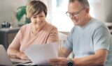 Et smilende par i middelalderen sidder ved et bord med en bærbar computer, en lommeregner og nogle papirer; de ser ud til at gennemgå pensionspapirer sammen i et lyst rum og diskuterer muligvis, hvordan de har investeret i Danmark med henblik på deres fremtid.