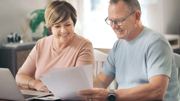 Et smilende par i middelalderen sidder ved et bord med en bærbar computer, en lommeregner og nogle papirer; de ser ud til at gennemgå pensionspapirer sammen i et lyst rum og diskuterer muligvis, hvordan de har investeret i Danmark med henblik på deres fremtid.