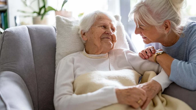 En ældre kvinde ligger på en sofa under et tæppe, mens en smilende kvinde, muligvis hendes datter, læner sig tæt ind mod hende og holder hende i hånden. De ser ud til at dele et varmt, kærligt øjeblik i en lys og hyggelig stue – måske taler de om muligheden for en flytning til et plejehjem og hvad det kunne betyde for deres fælles fremtid.