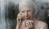 En ældre kvinde med hvidt hår holder en telefon op til øret, mens hun står ved et vindue med dug på ruden og ser bekymret eller urolig ud. Hun rører ved sit ansigt med den anden hånd og kigger ud, måske mens hun tænker over politiets opfordring til at advare de ældre mod svindel.