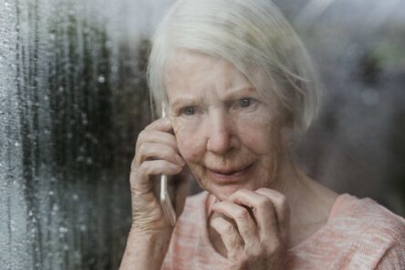 En ældre kvinde med hvidt hår holder en telefon op til øret, mens hun står ved et vindue med dug på ruden og ser bekymret eller urolig ud. Hun rører ved sit ansigt med den anden hånd og kigger ud, måske mens hun tænker over politiets opfordring til at advare de ældre mod svindel.
