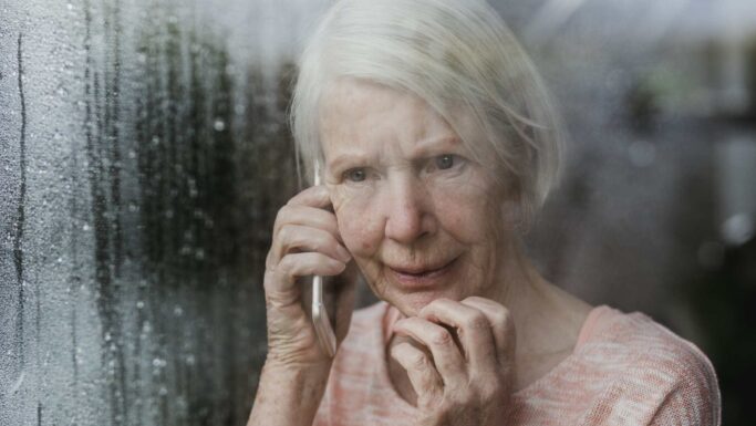 En ældre kvinde med hvidt hår holder en telefon op til øret, mens hun står ved et vindue med dug på ruden og ser bekymret eller urolig ud. Hun rører ved sit ansigt med den anden hånd og kigger ud, måske mens hun tænker over politiets opfordring til at advare de ældre mod svindel.