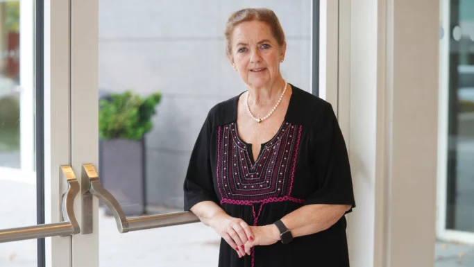En ældre kvinde står indenfor ved nogle glasdøre, iført en sort kjole med lyserødt broderi, en perlekæde og et ur, mens hun smiler blidt til kameraet. Udenfor kan man se grønne omgivelser gennem vinduet. Som medlem af Faglige Seniorer sætter hun pris på indsatsen for at beskytte ældre mod svindel i sit lokalsamfund.