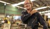 En ældre mand i arbejdsoveralls står i en fabrik og smiler med lukkede øjne, mens han holder sig for skulderen – muligvis på grund af ubehag eller smerter – en almindelig følge af hårdt arbejde for mange ældre. I den slørede baggrund ses maskiner og værktøj.