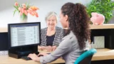 En kvinde sidder ved et skrivebord og arbejder på en computer, hvor der vises et regneark på skærmen, mens en ældre kvinde smiler til hende på den anden side af bordet. Der står blomster, en potteplante og en sparegris på bordet, mens de taler om bankgebyrer og kontooplysninger.