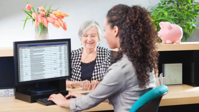 En kvinde sidder ved et skrivebord og arbejder på en computer, hvor der vises et regneark på skærmen, mens en ældre kvinde smiler til hende på den anden side af bordet. Der står blomster, en potteplante og en sparegris på bordet, mens de taler om bankgebyrer og kontooplysninger.