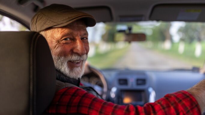 En ældre mand med gråt skæg og kasket smiler, mens han kører bil og kigger tilbage over skulderen. Sollyset stråler ind gennem bilen, og uden for forruden kan man se en landevej – et velkendt syn for mange pendlere på vej til arbejde, der måske endda tænker på skattefradrag, mens de kører deres daglige tur til og fra arbejde.