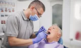 En tandlæge iført handsker og mundbind undersøger en ældre mandlig patients mund på en tandlægeklinik. Patienten sidder i en tandlægestol med en hagesmæk på, mens der i baggrunden ses tandlægeplakater, der understreger vigtigheden af regelmæssige besøg hos tandlægen gennem ti år.