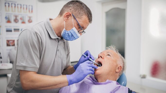 En tandlæge iført handsker og mundbind undersøger en ældre mandlig patients mund på en tandlægeklinik. Patienten sidder i en tandlægestol med en hagesmæk på, mens der i baggrunden ses tandlægeplakater, der understreger vigtigheden af regelmæssige besøg hos tandlægen gennem ti år.