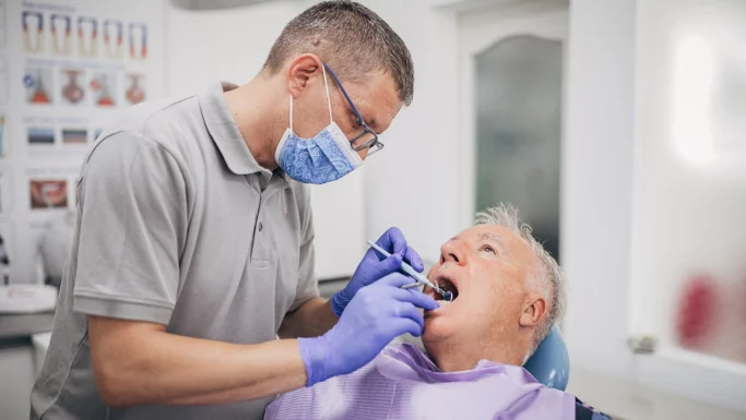 En tandlæge iført handsker og mundbind undersøger en ældre mandlig patients mund på en tandlægeklinik. Patienten sidder i en tandlægestol med en hagesmæk på, mens der i baggrunden ses tandlægeplakater, der understreger vigtigheden af regelmæssige besøg hos tandlægen gennem ti år.