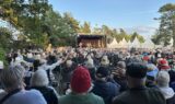 En stor menneskemængde sidder udendørs med udsigt til en scene, hvor musikere optræder ved Seniorfestivalen. Træer og hvide telte danner baggrunden under en klar himmel og bidrager til den festlige stemning. Mange mennesker løfter hænderne, fuldstændig opslugt af musikken og de spændende foredrag, der bydes på under hele arrangementet.