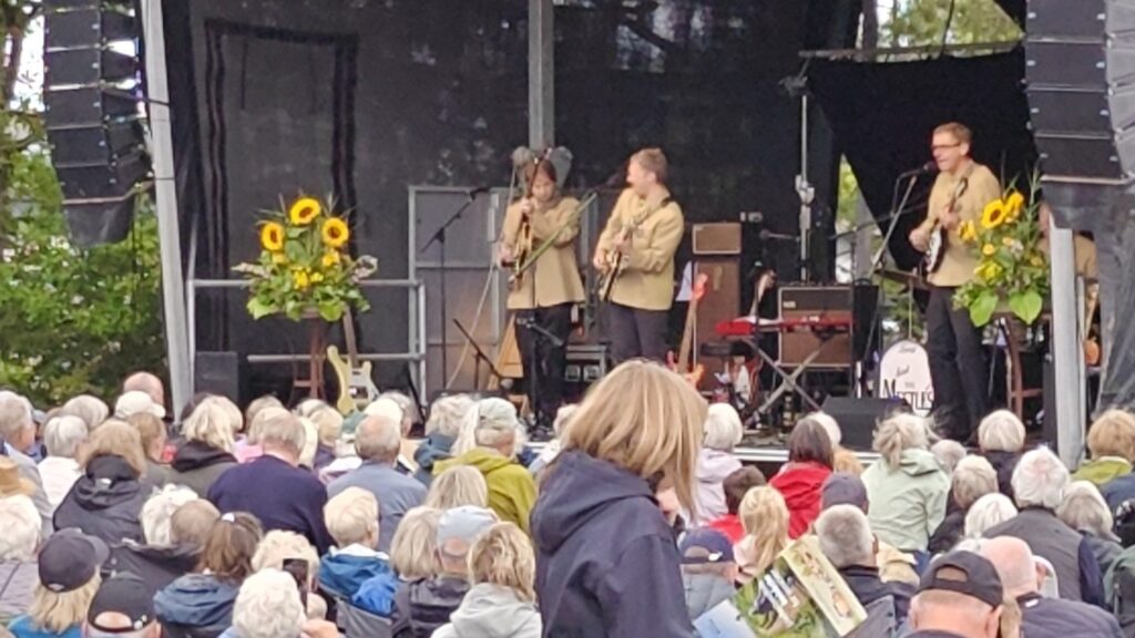 Et band i beige jakker optræder på en udendørs scene pyntet med solsikker ved Seniorfestivalen, mens et stort publikum, der sidder ned, følger optrædenen opmærksomt under en overskyet himmel, tager billeder af den livlige scene og spreder højt humør.