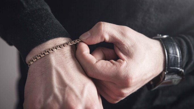 Et nærbillede af en persons hænder, der justerer et guldarmbånd om håndleddet. Personen har en mørk langærmet skjorte på og et sort armbåndsur på det andet håndled.