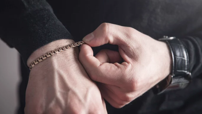 Et nærbillede af en persons hænder, der justerer et guldarmbånd om håndleddet. Personen har en mørk langærmet skjorte på og et sort armbåndsur på det andet håndled.