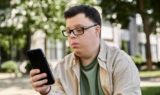 En ung mand med et udviklingshandicap, iført briller og afslappet tøj, sidder udendørs og kigger intenst på sin smartphone. I baggrunden ses træer og en bygning.