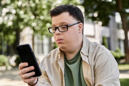 En ung mand med et udviklingshandicap, iført briller og afslappet tøj, sidder udendørs og kigger intenst på sin smartphone. I baggrunden ses træer og en bygning.