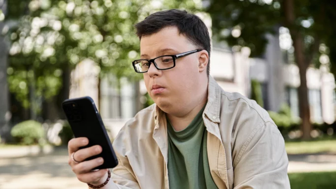 En ung mand med et udviklingshandicap, iført briller og afslappet tøj, sidder udendørs og kigger intenst på sin smartphone. I baggrunden ses træer og en bygning.
