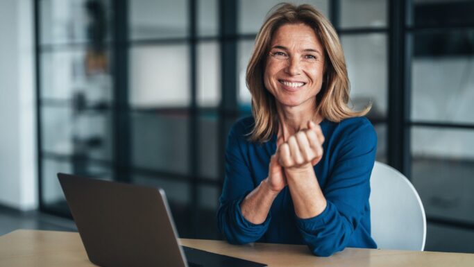 En smilende pensionist i en blå skjorte sidder ved et skrivebord med en bærbar computer og har hænderne foldet. Hun befinder sig i et moderne kontorlokale med glasvægge i baggrunden og er måske i gang med at drøfte tilskud eller økonomiske anliggender.
