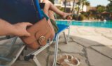 En person slapper af i en liggestol ved poolen, mens en anden i al hemmelighed begår et pungtyveri og tager en brun pung bag stolen. Der står sandaler på jorden, og i baggrunden ses en klarblå pool.
