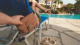 En person slapper af i en liggestol ved poolen, mens en anden i al hemmelighed begår et pungtyveri og tager en brun pung bag stolen. Der står sandaler på jorden, og i baggrunden ses en klarblå pool.