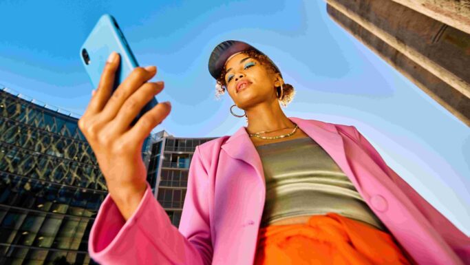 En person iført en lyserød blazer, orange bukser og en blå kasket står udendørs og kigger på sin smartphone – måske for at tjekke svindeladvarsler – med bygninger og en klar blå himmel i baggrunden, set fra et lavt vinkelperspektiv.