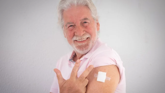 En smilende ældre mand i en lyserød skjorte peger på et plaster på sin overarm, hvilket viser, at han netop er blevet vaccineret mod lungebetændelse for at opnå en bredere beskyttelse. Han står foran en ensfarvet hvid baggrund.