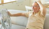 En ældre mand sidder på en sofa, holder sig for hovedet med den ene hånd og rækker ud mod en ventilator med den anden; han ser ud til at have det varmt og forsøger at køle sig ned i den varme i en lys og hyggelig stue.