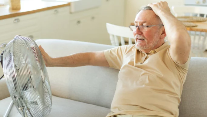 En ældre mand sidder på en sofa, holder sig for hovedet med den ene hånd og rækker ud mod en ventilator med den anden; han ser ud til at have det varmt og forsøger at køle sig ned i den varme i en lys og hyggelig stue.