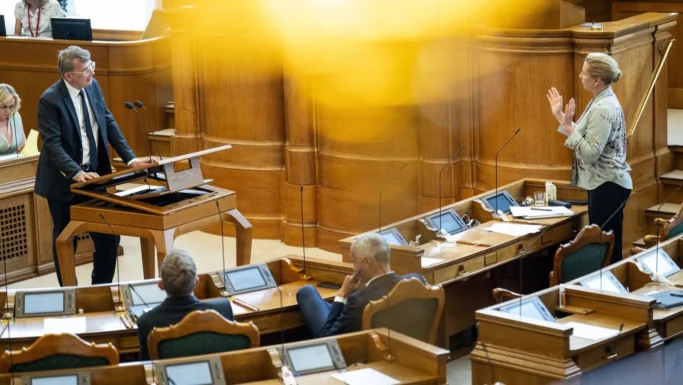 En kvinde står og gestikulerer, mens hun taler til en mand ved talerstolen i en formel parlamentssal med træpaneler, hvor debatter mellem de røde partier ofte udløser kraftig beskydning, mens flere personer lytter opmærksomt ved deres skriveborde med skærme.