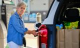 En ældre kvinde i en denimskjorte tanker diesel på sin bil ved en tankstation. Bilens bagagerum står åbent, og man kan se indkøbsposer fyldt med mad. I baggrunden ses en benzintank og en bygning.