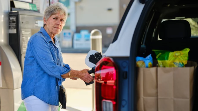 En ældre kvinde i en denimskjorte tanker diesel på sin bil ved en tankstation. Bilens bagagerum står åbent, og man kan se indkøbsposer fyldt med mad. I baggrunden ses en benzintank og en bygning.