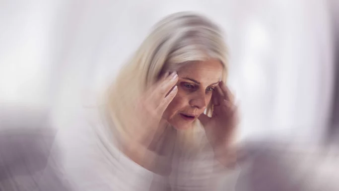 En ældre kvinde holder sig for tindingerne og ser bekymret ud, med en sløret, hvirvlende effekt omkring sig, der tyder på svimmelhed eller forvirring – tegn, der kan være udtryk for en psykisk krise, der kræver hjælp, f.eks. via en akuttelefon.