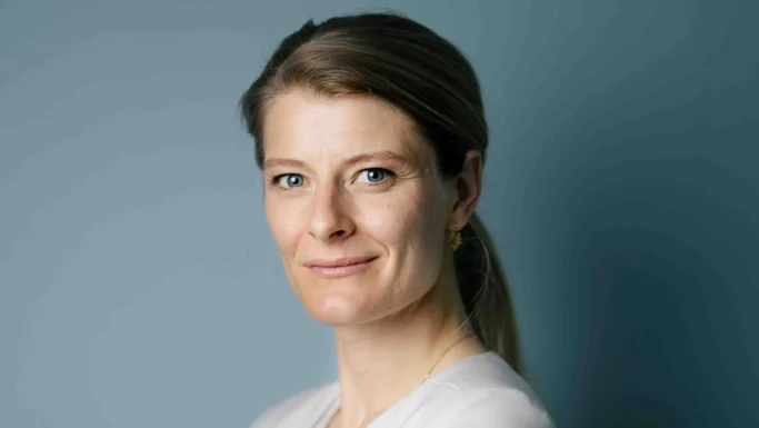En kvinde med lysebrunt hår, der er sat op, iført en lys bluse, ser ind i kameraet og smiler let på en ensfarvet blågrå baggrund – som symbol på de ældre, der udfordrer de fordomme, som nogle arbejdsgivere nærer.
