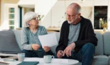 En ældre kvinde og en ældre mand sidder på en sofa og ser sammen på nogle papirer, mens de drøfter deres pensionsmuligheder. Kvinden holder et papir i hånden og taler med manden, mens de gennemgår deres planer. Der står kaffekopper og papirer på bordet foran dem i en lys, moderne stue.