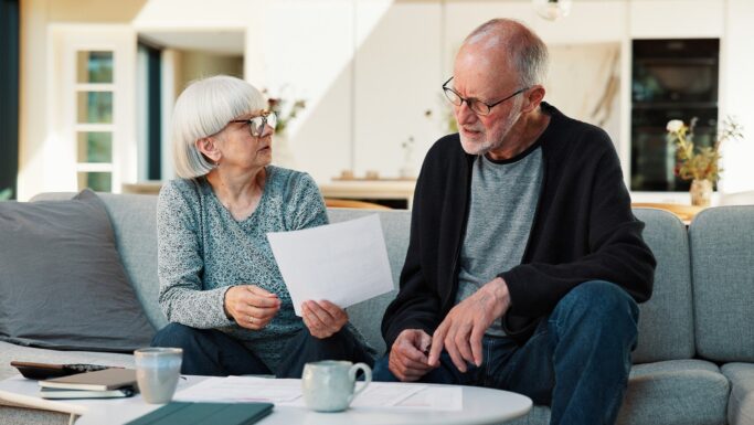 En ældre kvinde og en ældre mand sidder på en sofa og ser sammen på nogle papirer, mens de drøfter deres pensionsmuligheder. Kvinden holder et papir i hånden og taler med manden, mens de gennemgår deres planer. Der står kaffekopper og papirer på bordet foran dem i en lys, moderne stue.