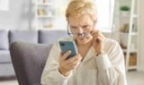 En pensionist med kort, blondt hår ser bekymret ud, mens vedkommende holder sine briller i hånden og stirrer på skærmen på en smartphone i en lyst stue.