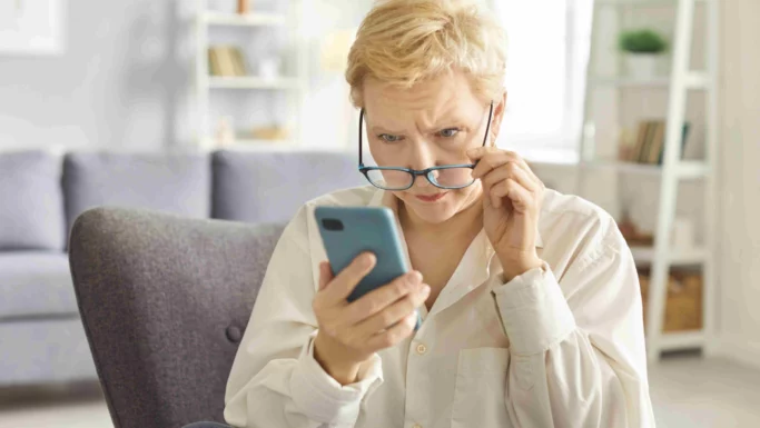 En pensionist med kort, blondt hår ser bekymret ud, mens vedkommende holder sine briller i hånden og stirrer på skærmen på en smartphone i en lyst stue.