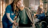 En smilende sygeplejerske støtter en ældre kvinde, der går med en rollator, og er dermed et levende eksempel på ånden bag »bedre trivsel« i ældreplejen. Den ældre kvinde læner kærligt hovedet mod sygeplejersken. I baggrunden sidder to andre ældre i kørestole og snakker sammen i et hyggeligt rum.
