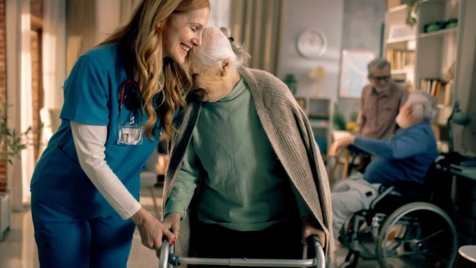 En smilende sygeplejerske støtter en ældre kvinde, der går med en rollator, og er dermed et levende eksempel på ånden bag »bedre trivsel« i ældreplejen. Den ældre kvinde læner kærligt hovedet mod sygeplejersken. I baggrunden sidder to andre ældre i kørestole og snakker sammen i et hyggeligt rum.