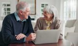En ældre mand og kvinde sidder ved et bord og smiler til hinanden, mens de med en åben bærbar computer foran sig i en lys stue diskuterer mulighederne for skattefradrag for seniorer.