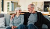 Et ældre ægtepar sidder på en sofa derhjemme og smiler varmt til hinanden, mens de taler om den nylige forhøjelse af folkepensionen. Manden har armen om kvindens skuldre, og begge virker tilfredse og trygge ved ændringerne i folkepensionen. I baggrunden ses en lys og moderne stue.