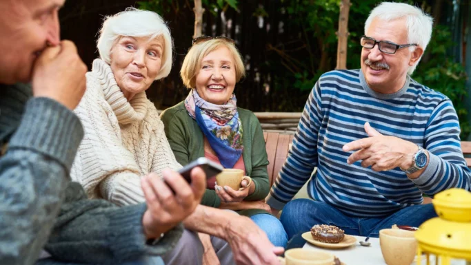 Fire ældre mennesker sidder sammen udendørs, smiler og griner, mens de holder kaffekopper i hånden. En af dem holder en telefon, og der står kager på bordet. De ser ud til at nyde en munter samtale i afslappede omgivelser, måske mens de udveksler historier om, hvordan de har forberedt sig på et nyt liv, når de går på pension.