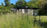 Et lille hus med glasvægge, der lokalt kaldes en »moderne bolig«, ligger omgivet af højt græs og frodig bevoksning under en klar, blå himmel.