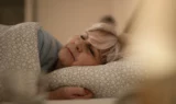 En ældre kvinde med gråt hår sover fredeligt på siden i sengen, med hovedet hvilende på en hvid pude med et cirkulært mønster og dækket af et matchende tæppe. Værelset fremstår blidt oplyst og roligt og giver et stemningsfuldt indblik i danskernes søvn.