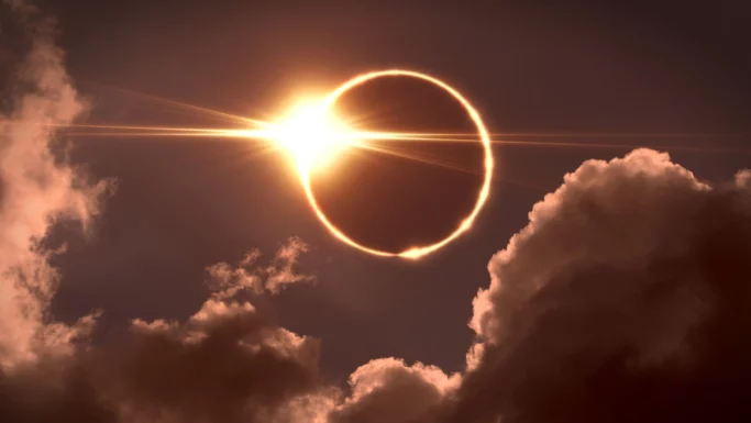 En solformørkelse, hvor solen danner en lys ring omkring månen, der er delvist skjult af skyer, på en mørk himmel.