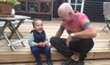Et smilende lillebarn og en ældre mand, sandsynligvis en bedsteforælder, sidder på en trætrappe udenfor og griner sammen. Ved siden af dem står et træbord med en potteplante. I baggrunden ses vinduer og en dør, hvilket skaber et varmt øjeblik, der hylder glæden ved børn på tværs af generationer – og de grænser, som kærlighed kan overvinde.